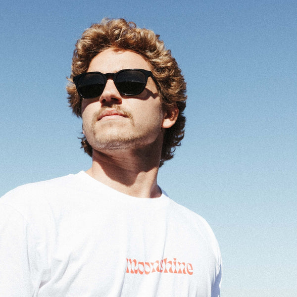 Man looking into the distance wearing black sunglasses and a white moonshine t shirt