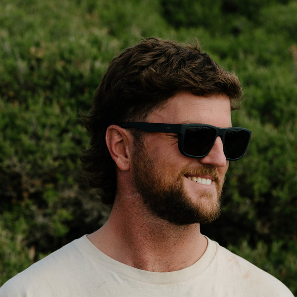 Man smiling wearing black sunglasses in front of a green bush
