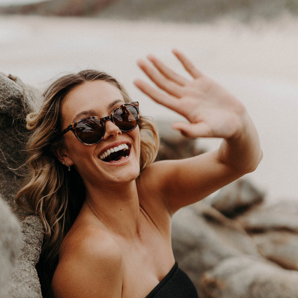 Girl smiling wearing sunglasses and waving her hand at the camera 
